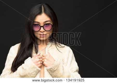 Picture of fashion asian model woman posing in sunglasses in studio. Attractive lady in white coat looking at camera.