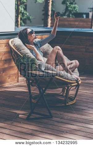 Capturing great moments. Beautiful young woman relaxing in a big comfortable chair on her outdoor house terrace and making selfie