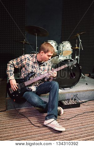 Bass guitar player sitting near drum set at music studio. Musician performing composition on bass guitar. Front view on playing bass guitar player at sound recording studio