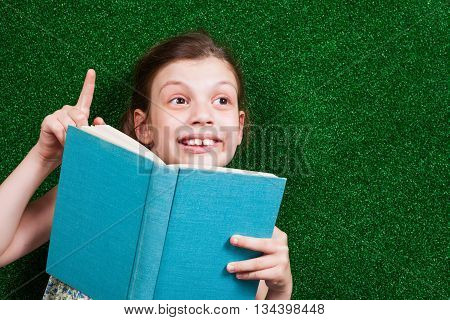 Little girl is lying on artificial grass. She is reading a book and having an idea.
