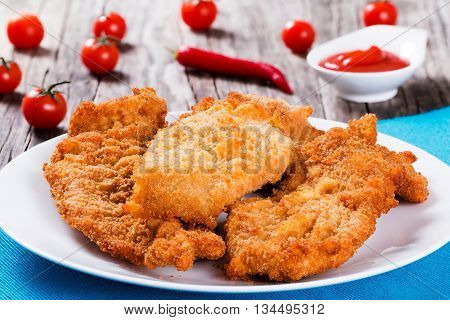 Bread Crumb Coated Fried Chicken breast on a white dish on a table mat with red chili pepper cherry tomatoes and ketchup in gravy boat on wooden background studio lights close-up