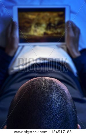 a young caucasian man lying in bed watches a movie or a series in streaming in his tablet computer, at night