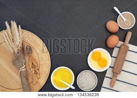 Bread baking ingredients and utensils with wheat sheaths and grain in a love spoon on grey slate background.
