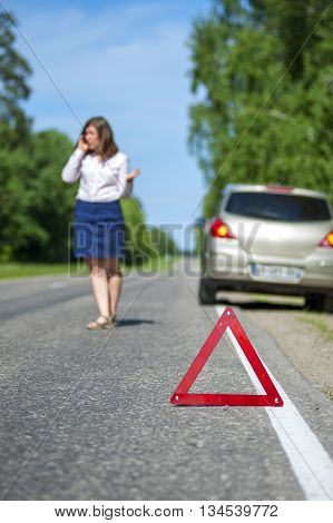 Woman calling to a car assistance after breakdown car on the roadside and red triangle warning sign. Focus on red sign