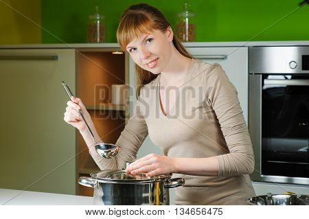 Girl on kitchen with a ladle. Smiling redhead woman with ladle cooking soup in home kitchen. Person removing lid from cooking pot. Beautiful young housewife looking at camera.