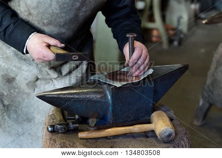 Old smith is chieseling a copper sheet in workshop, making pattern