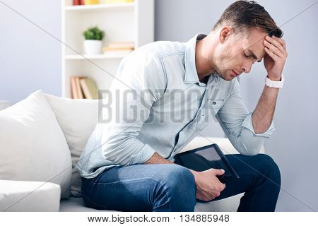 Terrible pain. Cheerless handsome sick man sitting on the couch and holding tablet while having a headache