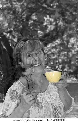 Middle aged woman with a happy look on her face posing with a flower and tea cup in front of trees. Selective color outdoor shot