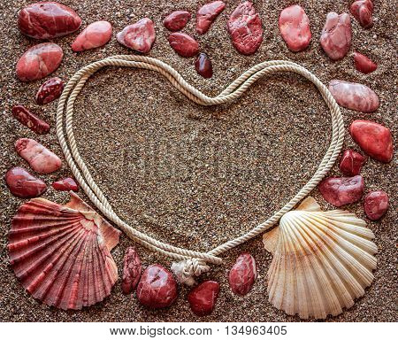 The concept of love. Around the frame in the shape of a heart arranged shells and stones red color. Sandy background.