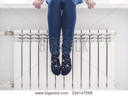 The Girl Sits On The Windowsill And Warms Her Feet In Pretty Socks At The Radiator, On A Cold Winter