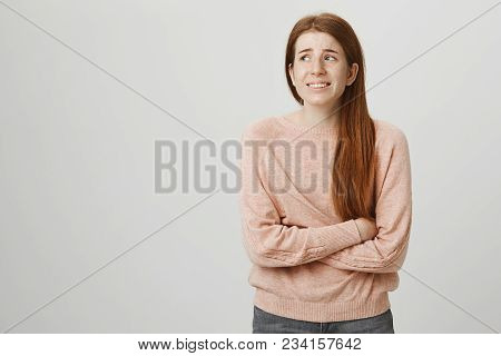 Indoor Portrait Of Cute Ginger Girl Standing With Crossed Hands On Belly, Feeling Awkward Or Sufferi