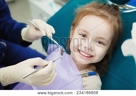 Child With Cute Smile Sits At Dentist Chair With Napkin On Chest And Doctor In Sterile Rubber Gloves