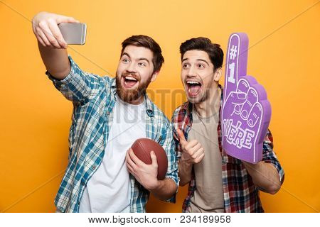 Portrait of a two excited young men taking a selfie while standing with rugby ball and foam glove isolated over yellow background