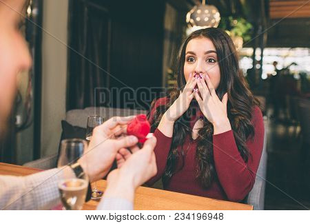 Good Shot Of Young Man Doing Propose To His Beloved Woman. He Is Showing Her A Box With Ring Inside 