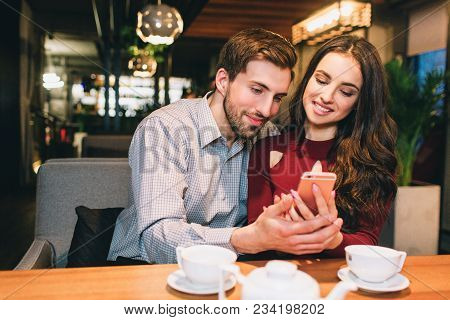 Young People Are Sitting Together In Cafe And Looking To The Phone. They Look Happy And Calm
