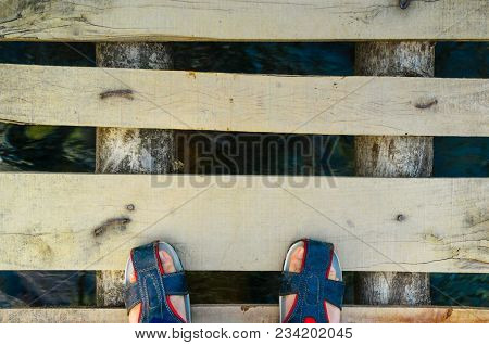 Feet In Fashionable Sandles Standing On A Wooden Plank Bridge With Water Flowing Under It. Shows The