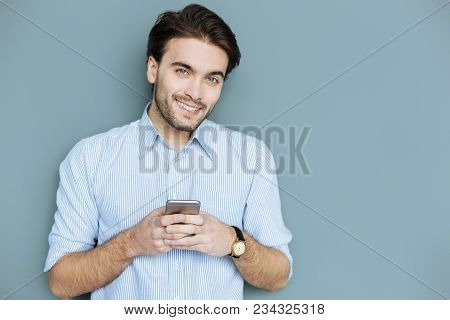 Modern Igen. Happy Nice Handsome Man Holding His Smartphone And Smiling To You While Typing A Messag