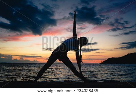 Full length side view of the silhouette of a fit woman, practicing the extended triangle yoga pose on a dreamy beach at twilight during summer vacation in Indonesia
