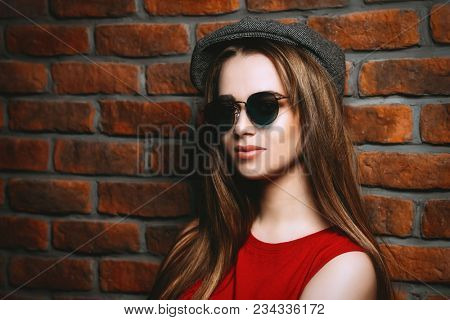 Attractive young woman in cap, sunglasses and red t-shirt stands by a brick wall. Beauty, fashion concept.