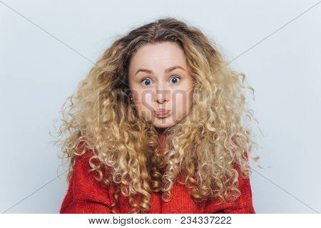 Fuuny Blonde Female With Curly Hair, Blows Cheeks And Stares At Camera, Reacts Actively On Unexpecte