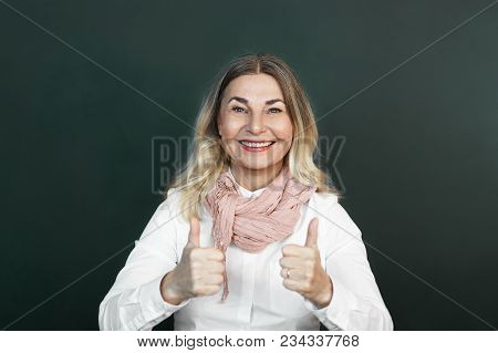 Happy Elderly Woman Wearing Elegant Mustard Scarf And White Shirt Smiling Broadly, Making Thumbs Up 