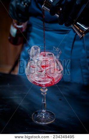 The Bartender Makes A Red Cocktail. Blue Dark Background. A Glass With Ice And Red Drink. Contrast P
