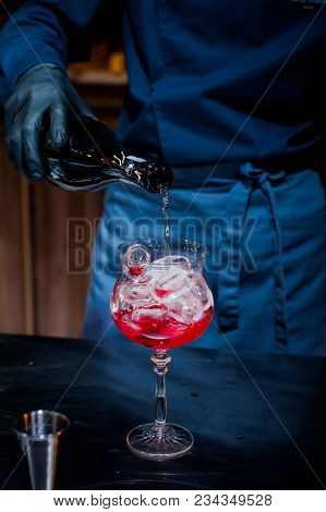 The Bartender Makes A Red Cocktail. Blue Dark Background. A Glass With Ice And Red Drink. Contrast P