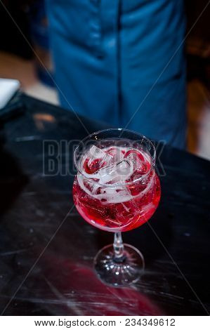 The Bartender Makes A Red Cocktail. Blue Dark Background. A Glass With Ice And Red Drink. Contrast P