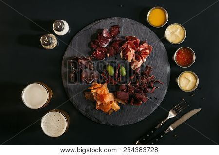 Top View Of Gourmet Assorted Meat On Slate Board, Various Sauces And Glasses Of Beer On Black