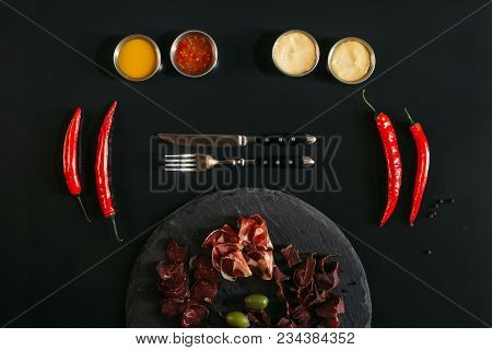 Gourmet Assorted Meat On Slate Board, Fork With Knife, Various Sauces And Chili Peppers On Black
