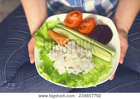 A Girl Holds A Plate With Rice And Vegetables. Healthy Eating Concept. Girl In Jeans And A Plaid Shi