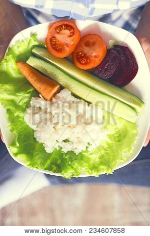 A Girl Holds A Plate With Rice And Vegetables. Healthy Eating Concept. Girl In Jeans And A Plaid Shi