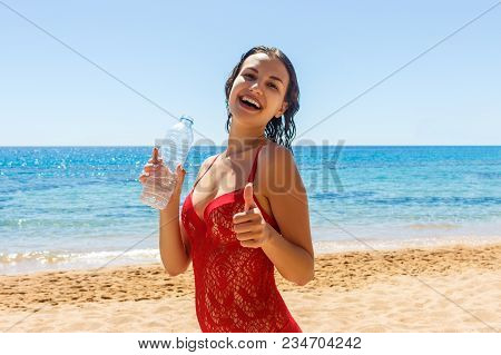 Happy Tanned Girl Holding A Water Bottle Gesturing Thumbs Up On The Beach.