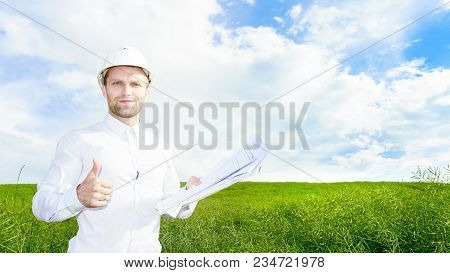 Geologist In White Helmet On Green Meadow With Drawings In Hands Holds His Thumb Up. Engineer Builde