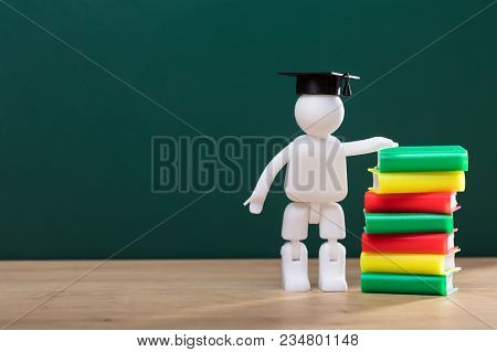 Human Figure With Graduation Hat Standing Beside Stacked Books On Wooden Desk