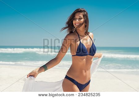 Beautiful girl with white fabric walking on the beach. Happy young woman relaxing at the beach and holding white scarf at wind in summer vacation. Latin tanned woman in blue bikini.