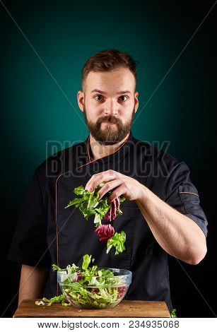 Portrait Of A Handsome Mischevious Male Chef Cook In Black Uniform Making Delicious Vegetable Salad 