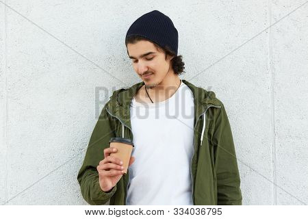 Image Of Handsome Sweet Young Boy Holding Papercup With Coffee, Looking At It Attentively, Wearing B