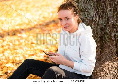 Woman In A White Hoodie Sweater With A Hood Sits On Ground In The Park And Holding A Cellphone In He