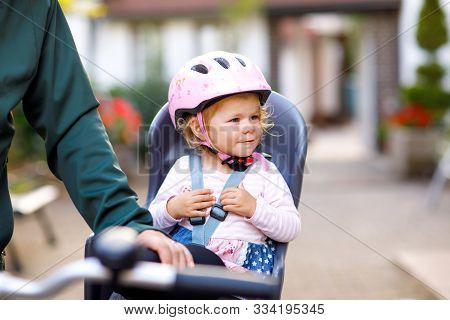 Portrait Of Little Toddler Girl With Security Helmet On The Head Sitting In Bike Seat And Her Father