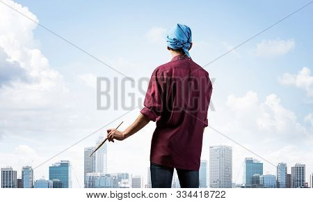 Young Male Painter Artist Holding Paintbrush. Back View Of Painter In Shirt And Bandana On Backgroun