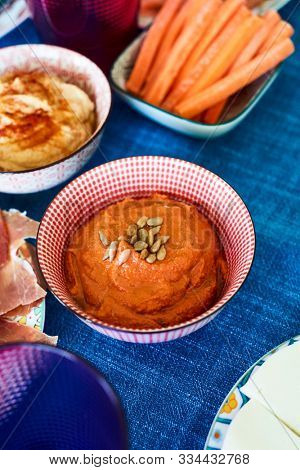 closeup of some plates and bowls with different appetizers, such as muhammara, hummus, strips of carrot, cheese or serrano ham, on a table set with a colorful blue tablecloth