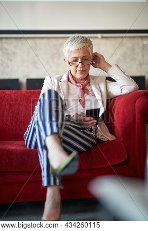 Elderly female boss sitting on the sofa during a break in a relaxed atmopshere at office and using a smartphone. Employees, office, work