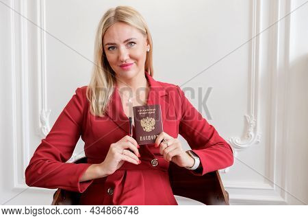 Attractive Blonde Woman In A Red Dress Holds A National Passport Of A Citizen Of The Russian Federat