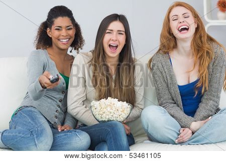 Portrait of cheerful young female friends with remote control and popcorn bowl on sofa at home