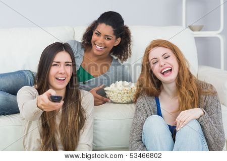 Portrait of cheerful young female friends with remote control and popcorn bowl on sofa at home