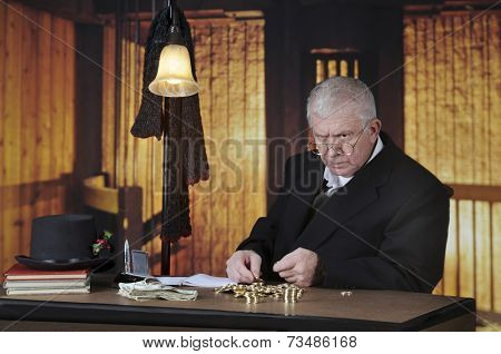 A grumpy old miser sitting at his desk counting gold coins by a stack of big bills.