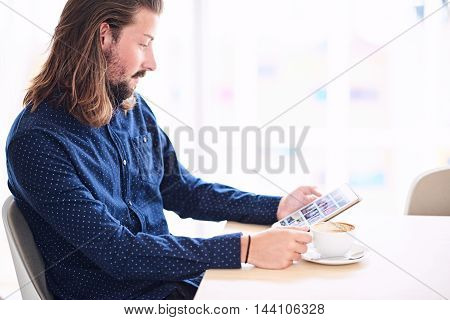 Young caucasian man sitting at a table next to a large window while holding a tablet in his hands and a cappaccino on the table in front of him.