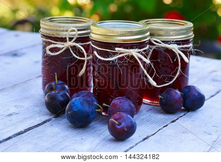 Bottles of homemade plum jam and some plums on an outside picnic table.