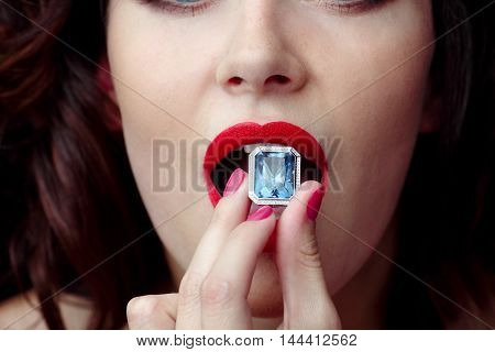 Female mouth with red lipstick puts a hand to the ring with a big sapphire diamond to eat, swallow, close up